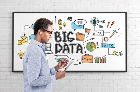 Side view of pensive young African American man in glasses making notes standing near whiteboard with colorful big data sketch drawn on it.の写真素材