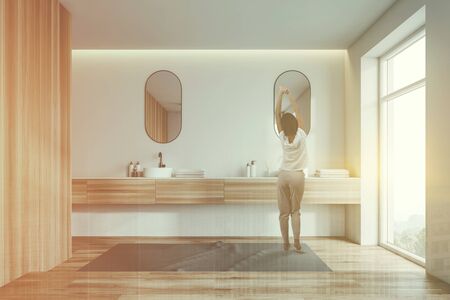 Rear view of woman in pajamas standing in luxury white and wooden bathroom interior with wooden floor, large window and double sink with oblong mirrors above it. Toned image double exposureの写真素材