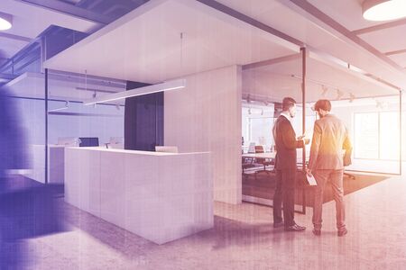 Two businessmen discussing documents in office hall with white reception table and meeting room in background. Toned image double exposure blurredの写真素材