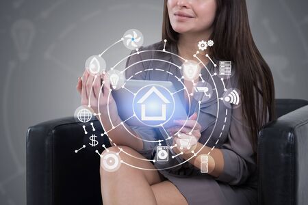 Unrecognizable woman with long dark hair sitting in armchair with tablet computer in gray room with double exposure of smart home interface. Concept of automation. Toned imageの写真素材