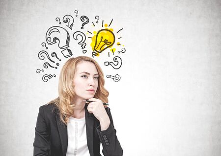 Portrait of thoughtful young businesswoman with red hair wearing suit and looking forward sitting near concrete wall with question marks and light bulb on it. Mock upの写真素材