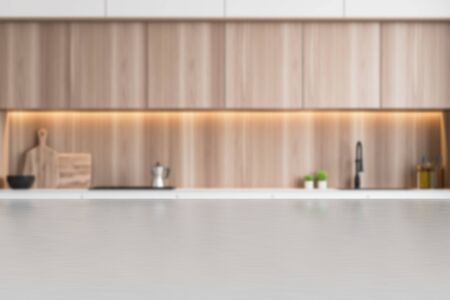 Blurred interior of minimalist kitchen with white and wooden walls, white countertops with built in sink and cooker and wooden cupboards. Table for your product in foreground. 3d renderingの写真素材