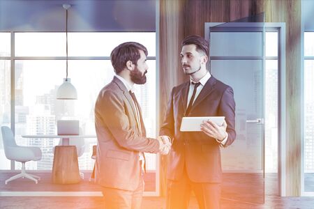 Two young businessmen shaking hands in modern coworking office with wooden walls, concrete floor and round table with laptop and white chairs. Toned imageの写真素材