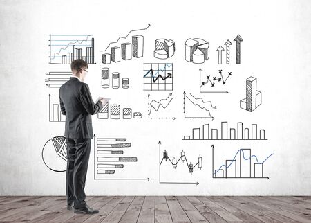Side view of young caucasian businessman in glasses taking notes while looking at graphs drawn on concrete wall. Concept of market analysisの写真素材
