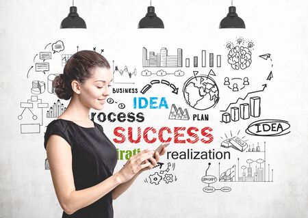 Smiling young woman in black dress looking at her smartphone standing near concrete wall with colorful business success sketch drawn on itの写真素材