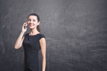 Portrait of young smiling businesswoman in black dress talking on smartphone and looking away...