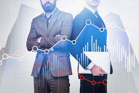 Businessman with laptop and his confident bearded colleague standing over cityscape background with double exposure of graphs. Concept of trading. Toned imageの写真素材