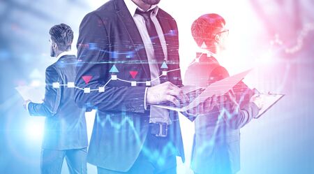 Businessman with laptop and his colleagues with documents standing together over blurred background with double exposure of graphs. Concept of teamwork and stock market. Toned imageの写真素材