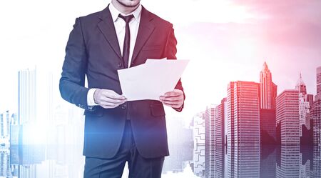 Unrecognizable young businessman in dark suit reading documents standing over modern cityscape background. Concept of paperwork. Toned imageの写真素材