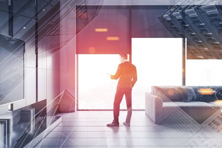 Man in suit standing in modern living room interior with white and gray walls, large windows, gray sofa and TV set. Toned image double exposureの写真素材