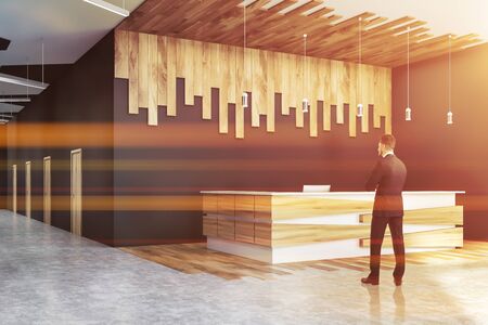 Businessman standing in office hall with gray and wooden walls, concrete floor and white and wooden reception desk. Toned imageの写真素材