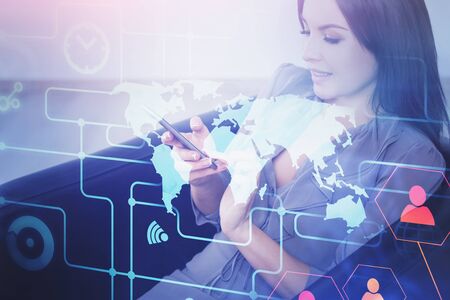 Smiling young businesswoman sitting in leather armchair with smartphone and double exposure of social network hologram and world map. Concept of social interaction in business. Toned imageの写真素材