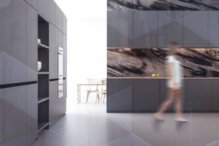 Young woman walking in stylish kitchen with black marble walls, concrete floor, gray countertops and...