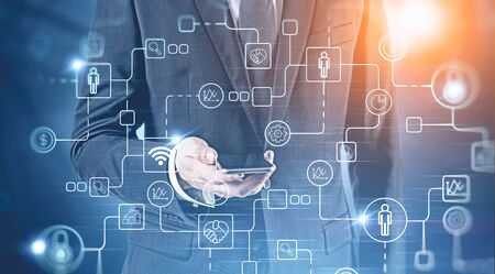 Unrecognizable businessman in dark suit looking at smartphone over blurred background with double exposure of digital icons. Concept of hi tech and corporate lifestyle. Toned imageの写真素材