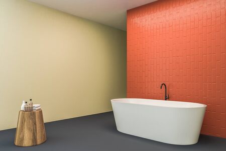Corner of bright bathroom with beige and orange walls, concrete floor, white bathtub and wooden table with towels and bottles. 3d renderingの写真素材