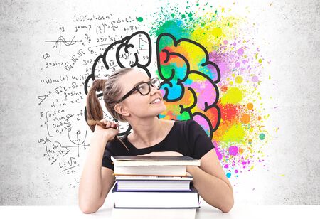 Smiling blonde woman in glasses sitting at table with stack of books near concrete wall with brain sketch and formulas. Concept of education and scienceの写真素材