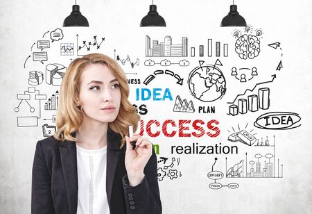 Thoughtful young woman in suit with ginger hair standing near concrete wall with business plan sketch drawn on it. Concept of business strategyの写真素材
