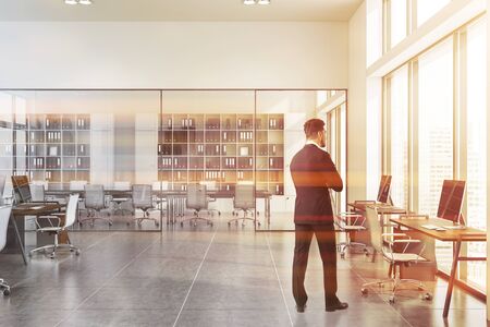Rear view of businessman standing in stylish office interior with white walls, tiled floor, open space area and conference room in background. Toned imageの写真素材