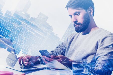 Young software developer in casual clothes working with laptop and smartphone in city with double...