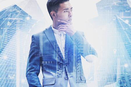 Thoughtful Asian businessman in suit standing in modern city with double exposure of HUD network interface. Concept of software development and smart city. Toned imageの写真素材