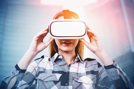 Astonished young blonde woman in checkered shirt and VR glasses over blurred city background....