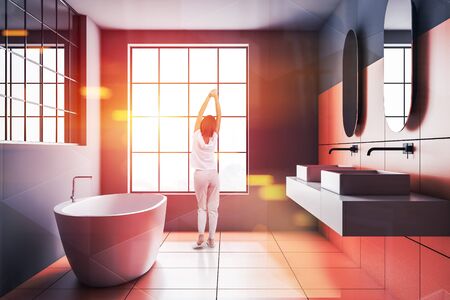 Rear view of woman in pajamas standing in modern bathroom with gray and orange tile walls, big window, double sink and comfortable white bathtub. Toned image double exposureの写真素材