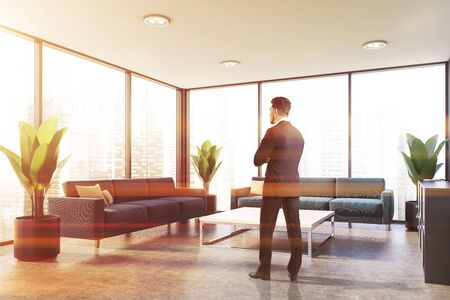 Young businessman standing in panoramic office waiting room with blue sofas and square coffee table. Toned imageの写真素材
