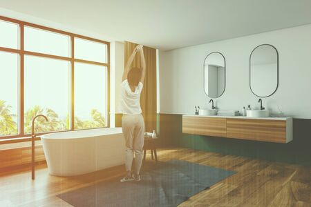 Young woman standing in modern bathroom corner with white and green walls, wooden floor with carpet, comfortable white bathtub and double sink. Toned image double exposureの写真素材