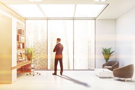 Young businessman standing in modern office with white and wooden walls, computer table with folders above it and lounge area with gray armchairs. Toned imageの写真素材