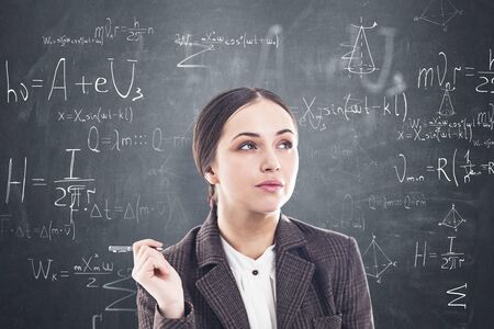 Thoughtful young woman with dark hair and pen standing near blackboard with formulas written on it....