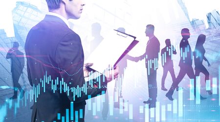 Rear view of businessman with clipboard standing in modern city with business people silhouettes and double exposure of digital graphs. Concept of management and stock market. Toned imageの写真素材