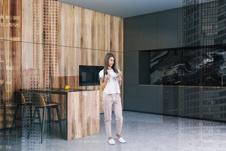 Young woman standing in modern kitchen interior with dark countertops, black marble walls and wooden table with bar stools. Toned image double exposureの写真素材