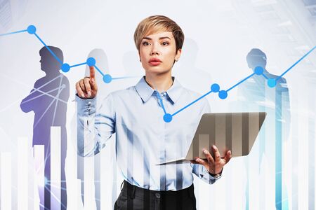Young businesswoman with laptop working with digital graph in city with double exposure of business people silhouettes. Concept of teamwork and stock market. Toned imageの写真素材