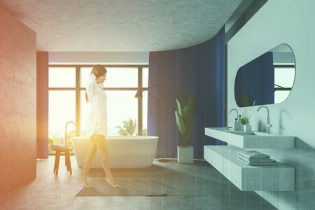 Young woman walking in stylish sunlit bathroom with blue curtains, double sink and white bathtub. Toned image double exposureの写真素材