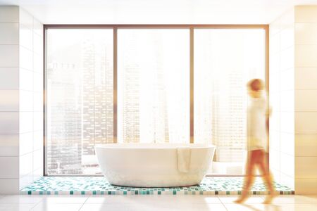 Blurry young woman walking in panoramic bathroom with cityscape, white tile walls, blue mosaic floor and comfortable white bathtub. Toned imageの写真素材