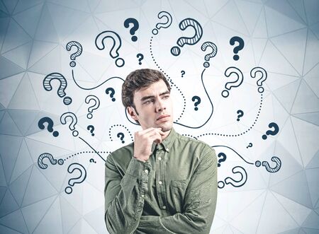 Thoughtful young man in green shirt standing near gray wall with question marks drawn on it. Concept of problem solving and curiosityの写真素材