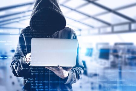 Young unrecognizable hacker in black hoodie using laptop over blurry office background with double exposure of lines of code. Concept of identity theft and cyber security. Toned imageの写真素材