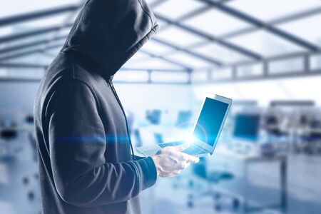 Young hacker standing with laptop in blurry office. Concept of cyber crime, online security and identity theft. Toned imageの写真素材