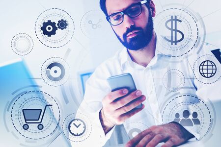 Bearded businessman in glasses holding smartphone in blurry office with double exposure of online shopping interface. Toned imageの写真素材