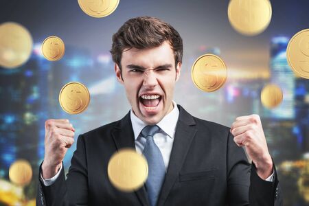 Happy young businessman screaming with joy standing under dollar rain in blurry night city. Concept of business successの写真素材