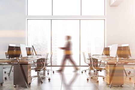 Blurry young businessman walking in modern open space office with white walls, concrete floor, big window with cityscape and wooden computer desks. Toned imageの写真素材