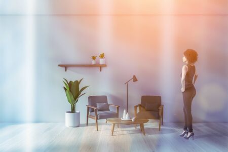 Young African American woman standing in modern living room with white walls, white wooden floor, two grey armchairs near coffee table and shelf with plants. Toned imageの写真素材