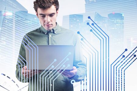Serious young programmer using laptop in modern city with double exposure of circuit interface....