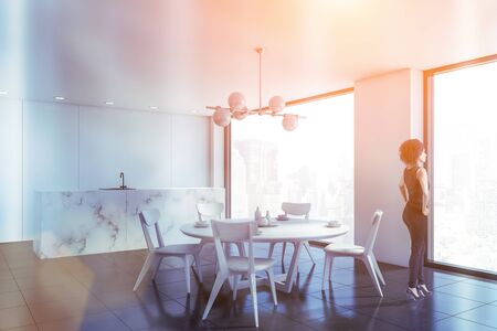Young African American woman standing in luxury kitchen with white walls, marble countertop and round dining table with chairs. Toned imageの写真素材