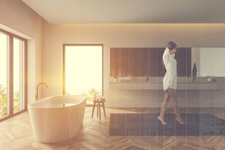 Young woman in nightgown walking in loft bathroom with wooden and white walls, wooden floor, comfortable tub and gray double sink. Toned image double exposureの写真素材