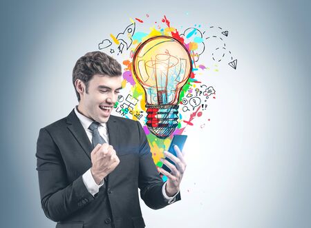 Happy successful young businessman with smartphone standing near gray wall with colorful business idea sketch drawn on it. Concept of new project launchの写真素材