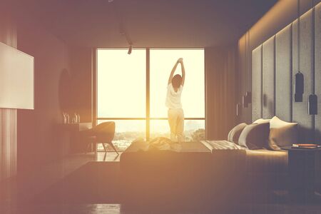 Young woman in pajamas standing in sunlit bedroom with gray walls, king size bed, makeup table with round mirror and armchair and TV on the wall. Toned image double exposureの写真素材