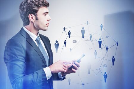 Handsome young businessman using smartphone in blurry city with double exposure of social network interface. Concept of job search and communication. Toned imageの写真素材