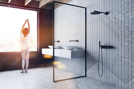 Young woman in pajamas standing in sunlit bathroom with tiled and gray walls, concrete floor, double sink on white countertop and shower stall. Toned image double exposureの写真素材