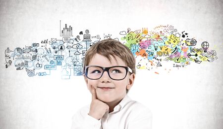 Smiling adorable little boy in glasses standing near concrete wall with creative business plan sketch drawn on it.の写真素材
